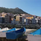 Small blue and white fishing boats docked at the harbor in A Guarda, Spain, with colorful hillside buildings in the background on a sunny day.
