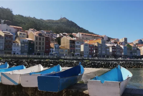 Small blue and white fishing boats docked at the harbor in A Guarda, Spain, with colorful hillside buildings in the background on a sunny day.