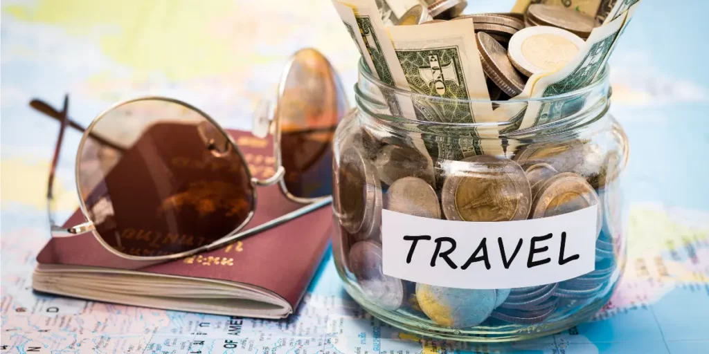 A travel savings jar filled with coins and US dollars, placed next to a passport and sunglasses on a world map, symbolizing Camino budget planning.