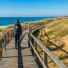 Solo traveler on the Portuguese Coastal Way walking a wooden boardwalk beside Atlantic dunes, wearing a backpack marked with a scallop shell.