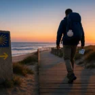 Camino de Santiago pilgrim at dawn on the Portuguese Coastal Way boardwalk, scallop shell on backpack, granite waymark and Atlantic surf in view.